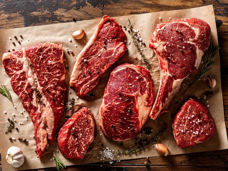 Ribeye, strip, and filet steaks laid out on butcher paper with herbs and peppercorns.