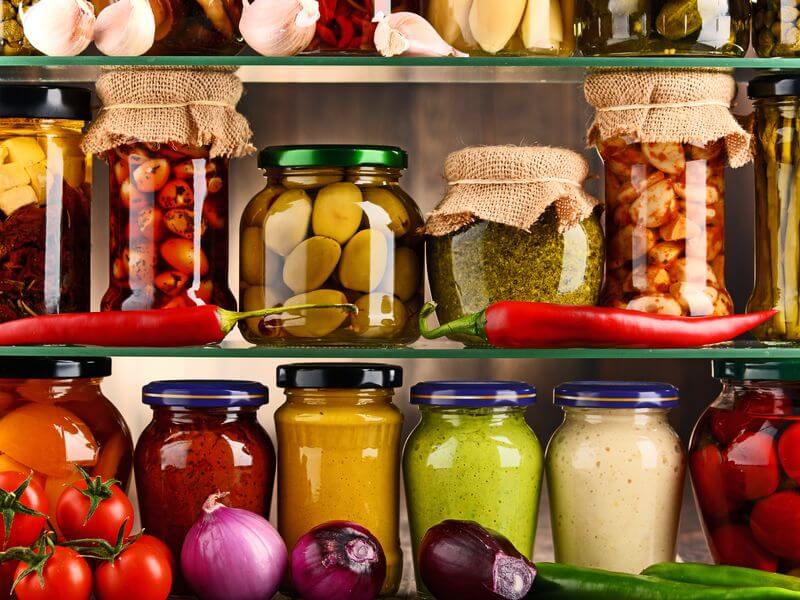Shelves of jarred preserves, pickles, and sauces above baskets of fresh produce.